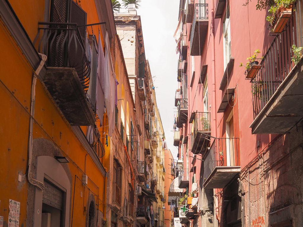 A narrow street with ochre and pink buildings and balconies in historic centre of Naples. Copyright@2019 mapandfamily.com 