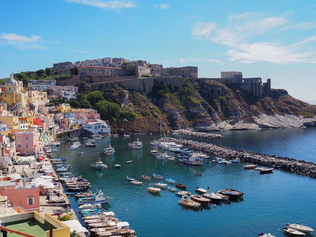 Pastel coloured houses by harbour with boats and dark buildings on headland. 