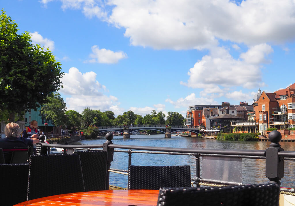 A table and chairs on outdoor terrace overlooking River Thames with Eton bridge in background. Copyright@2023 mapandfamily.com 