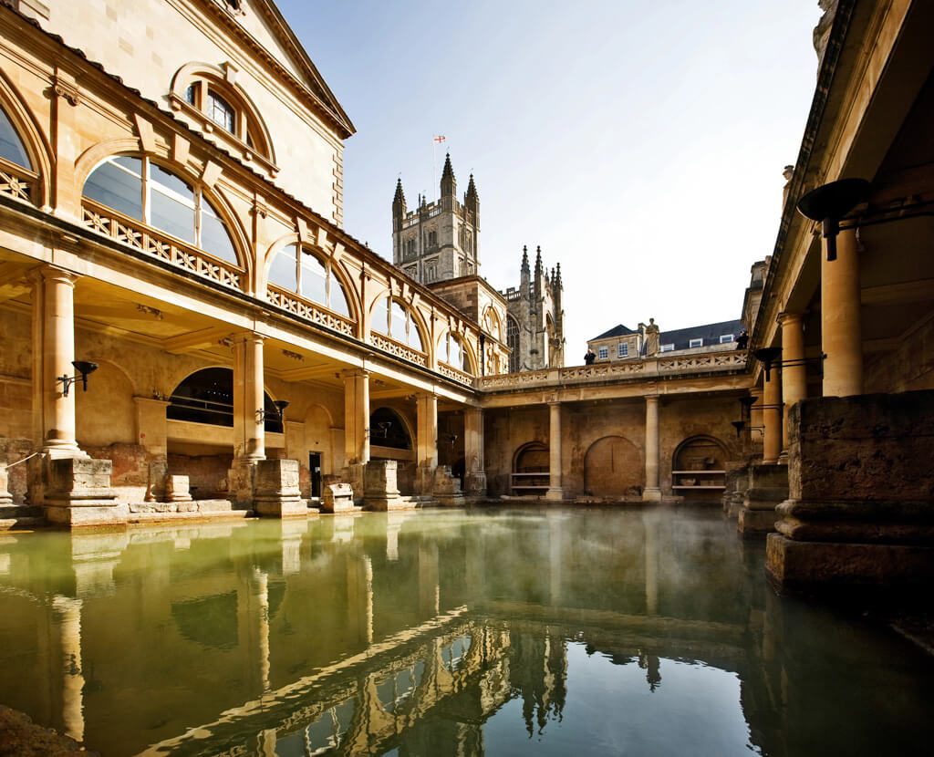 Ancient Roman baths with stone colums reflected in green water in Bath. DepositPhotos