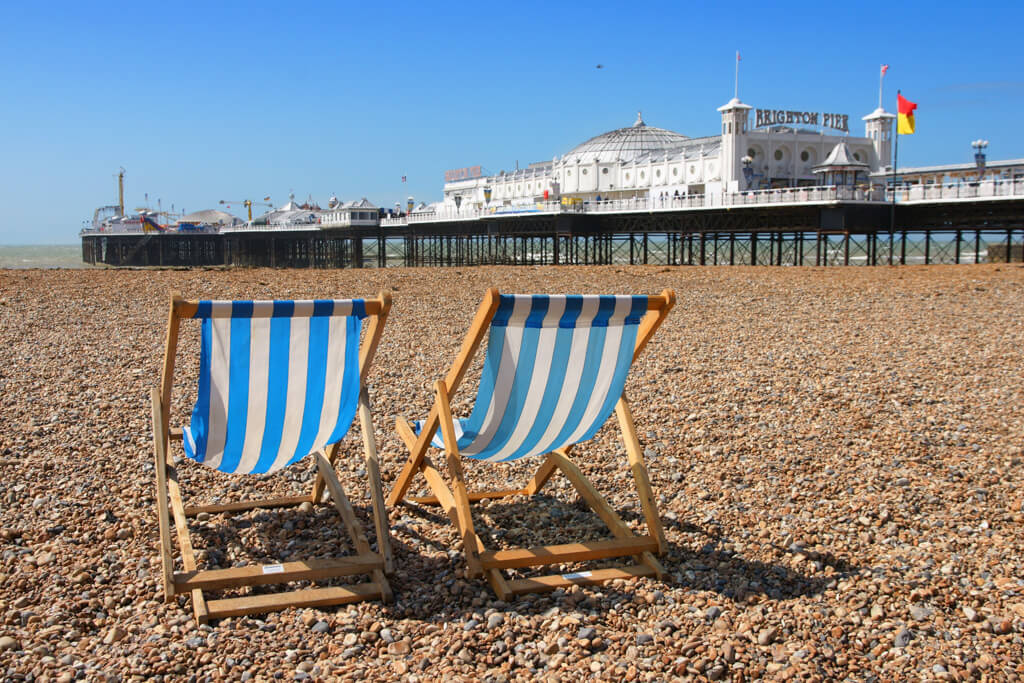 Blue and white striped deckchairs on pebble beach beside white painted pier in Brighton. DepositPhotos