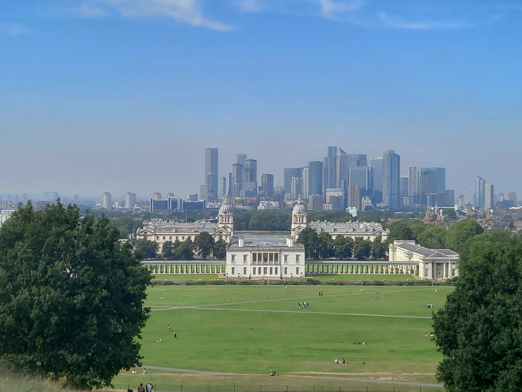 A view across Greenwich park to the grand and formal complex of historic buildings. In the background are the skyscrapers of Canary Wharf. Coopyright@2024 mapandfamily.com