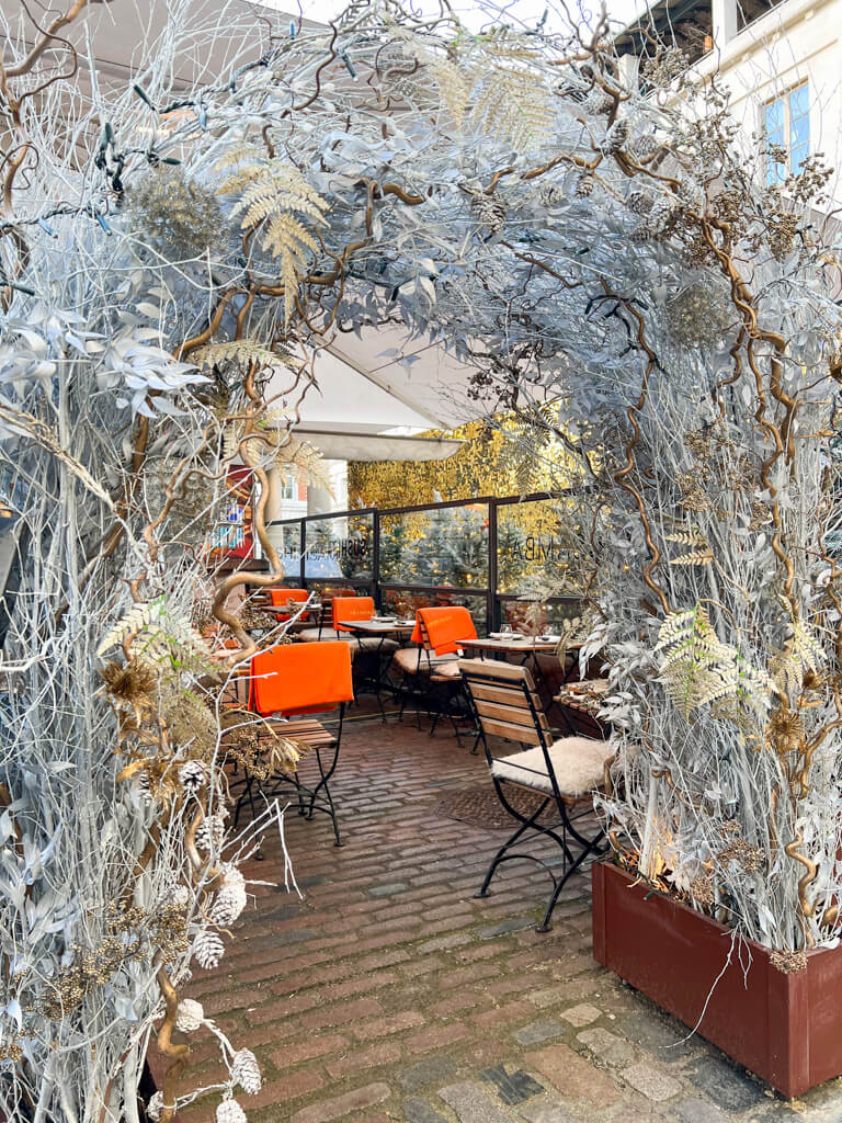 Gold and silver twiggy archway leading to outdoor terrace with tables, chairs and orange blankets. Copyright@2024 mapandfamily.com