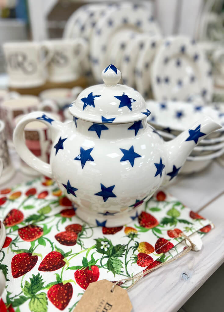 A white teapot decorated with blue stars on a strawberry-patterned teacloth. Copyright@2025 NancyRoberts 